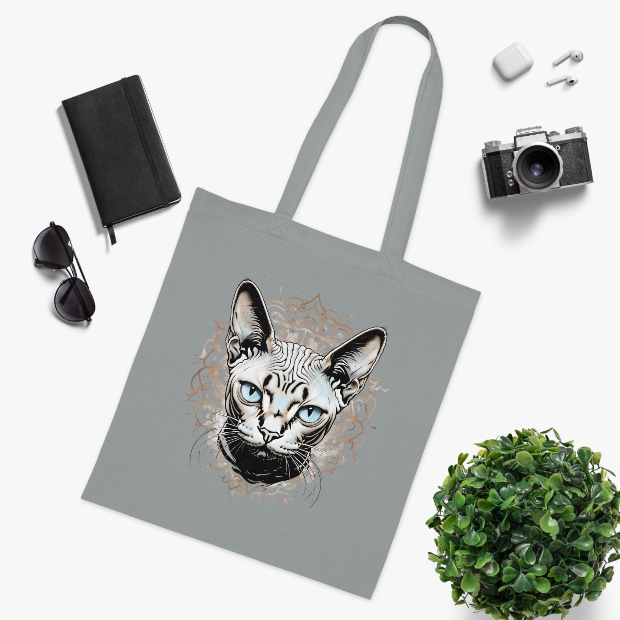 Gray tote bag with a cat design surrounded by a camera, notebook, sunglasses, and plant on a white background