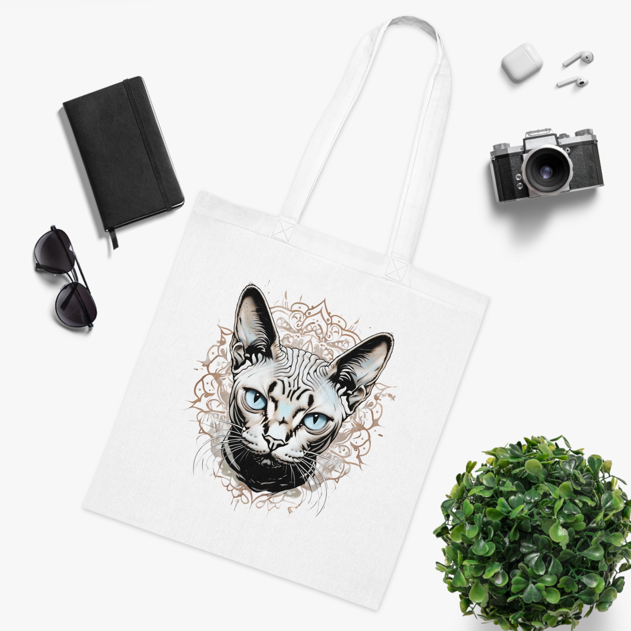 White tote bag with a cat design, surrounded by sunglasses, camera, notebook, and plant on a white background