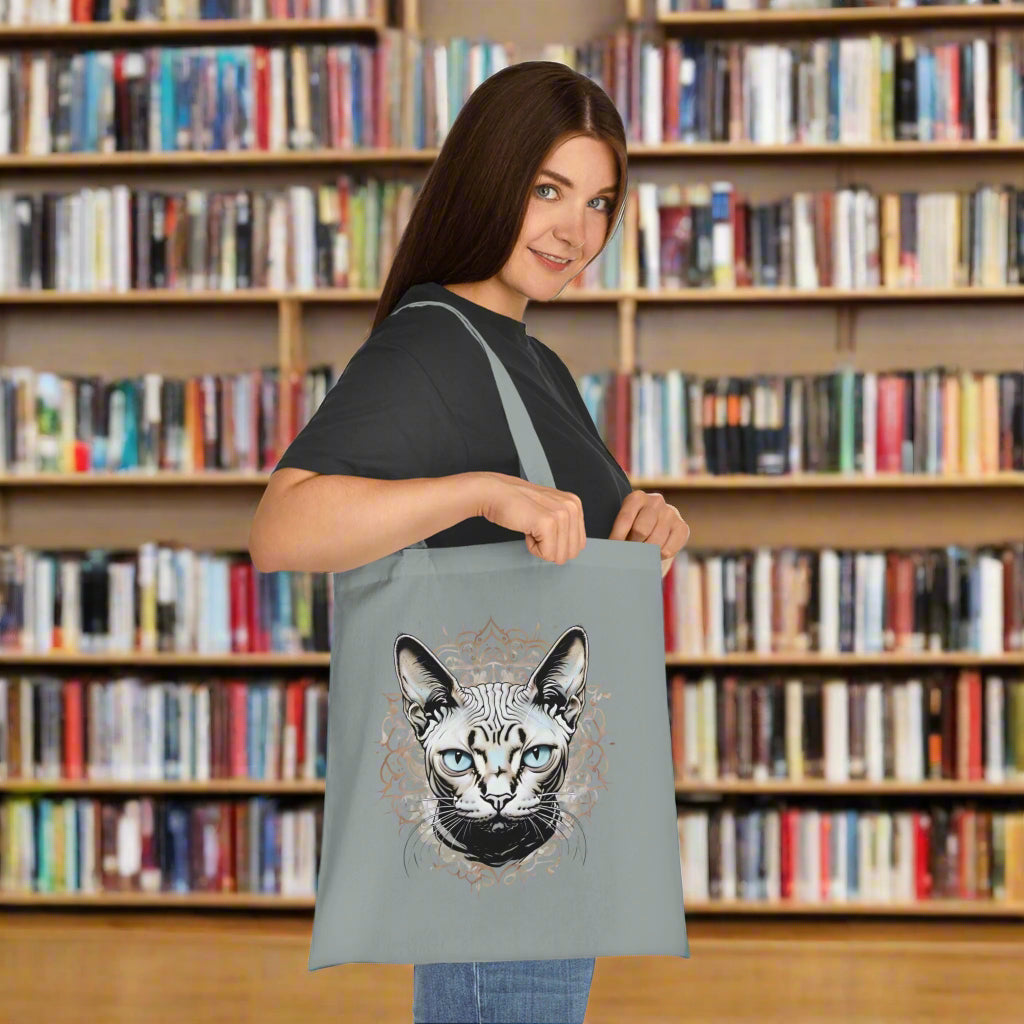 Person holding a grey tote bag with a sphynx cat design in a library