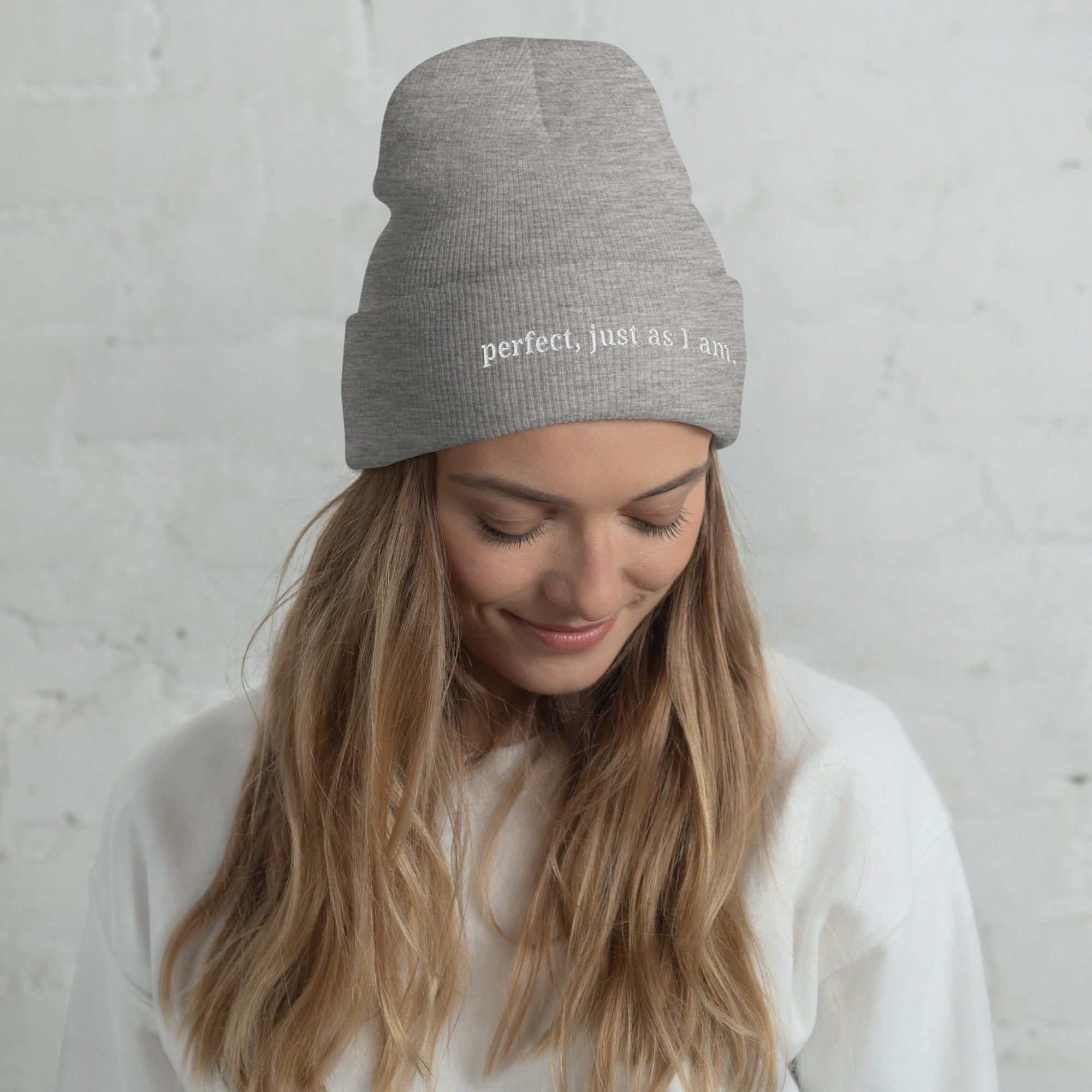 Woman wearing a grey beanie with 'perfect, just as I am' text embroidery against a light background