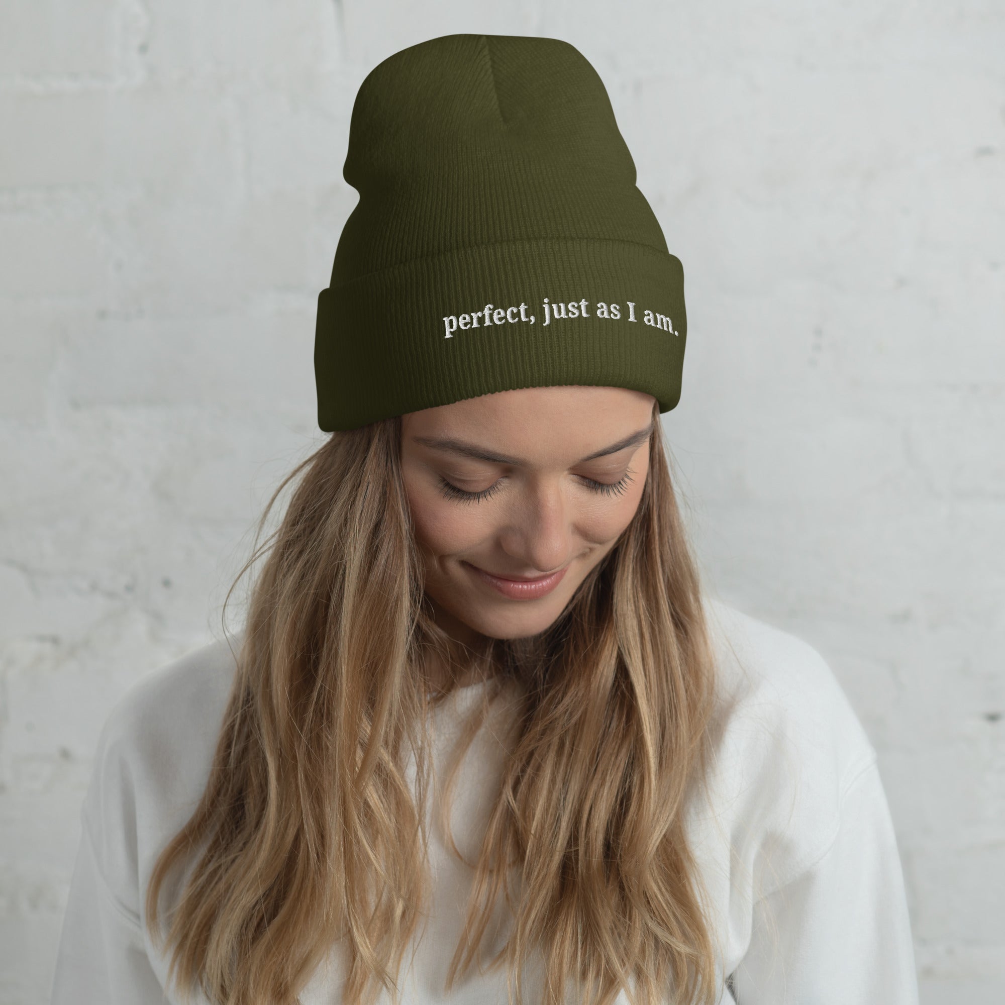 Woman wearing a green beanie with 'perfect, just as I am' text embroidery against a light grey background