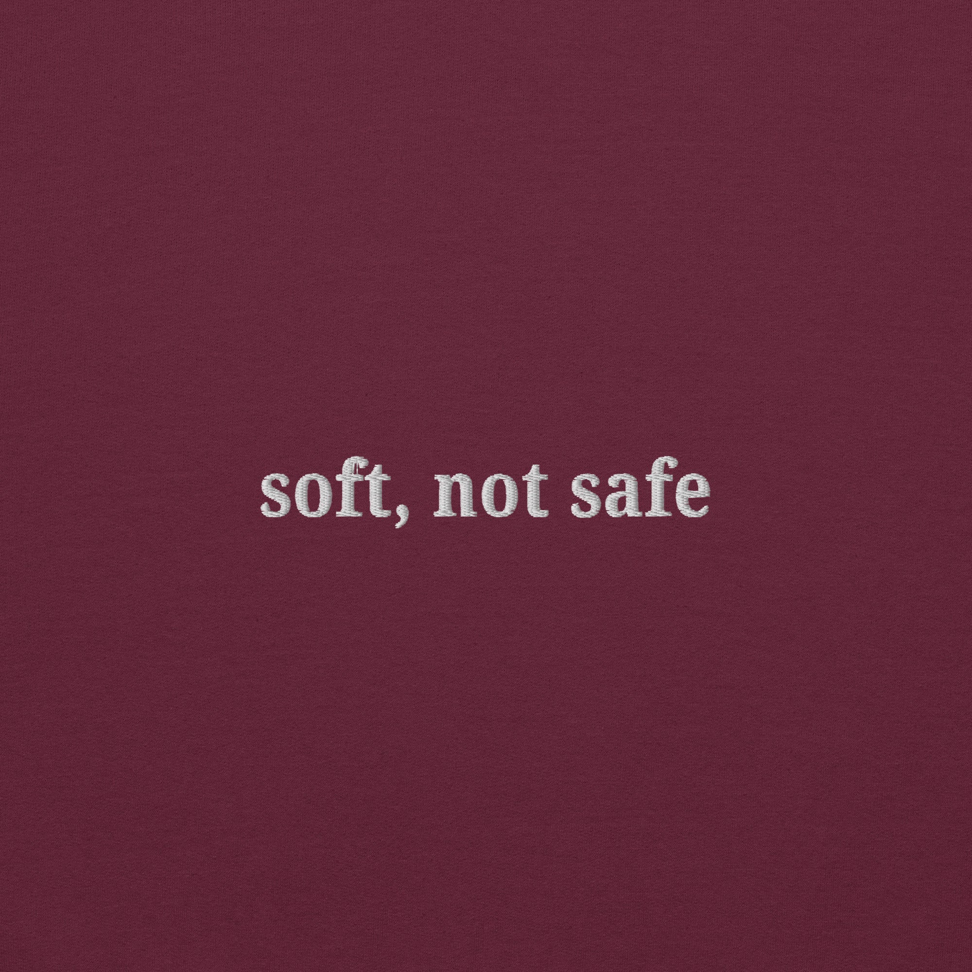 Close up of white thread embroidery reading Soft, Not Safe on maroon Gildan unisex sweatshirt
