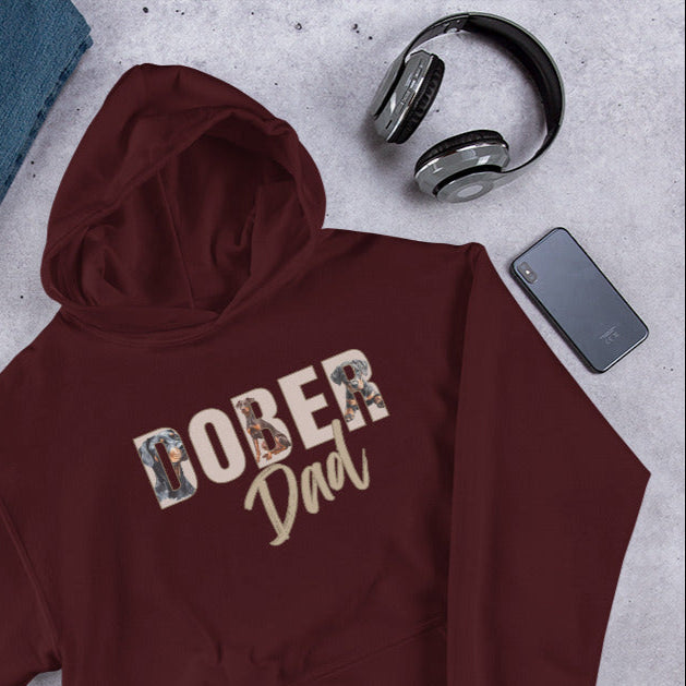 Maroon hoodie with 'DOBER Dad' text, surrounded by jeans, shoes, camera, and accessories on a gray surface.