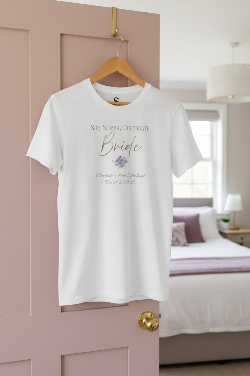 Personalised sober bride t-shirt in white with metallic-effect text and mauve accent details, hanging on a door