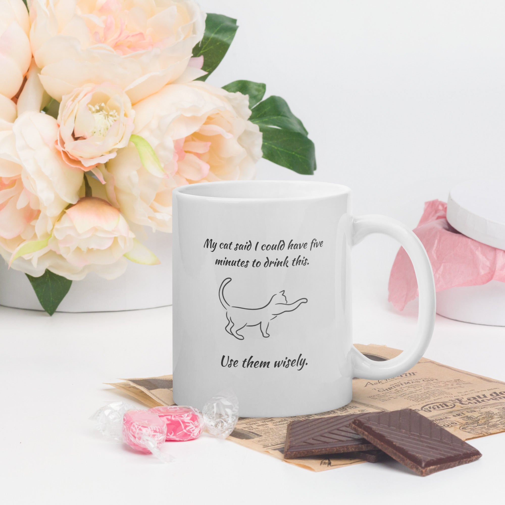 White mug with humorous text and a cat illustration on a table with flowers and chocolate.