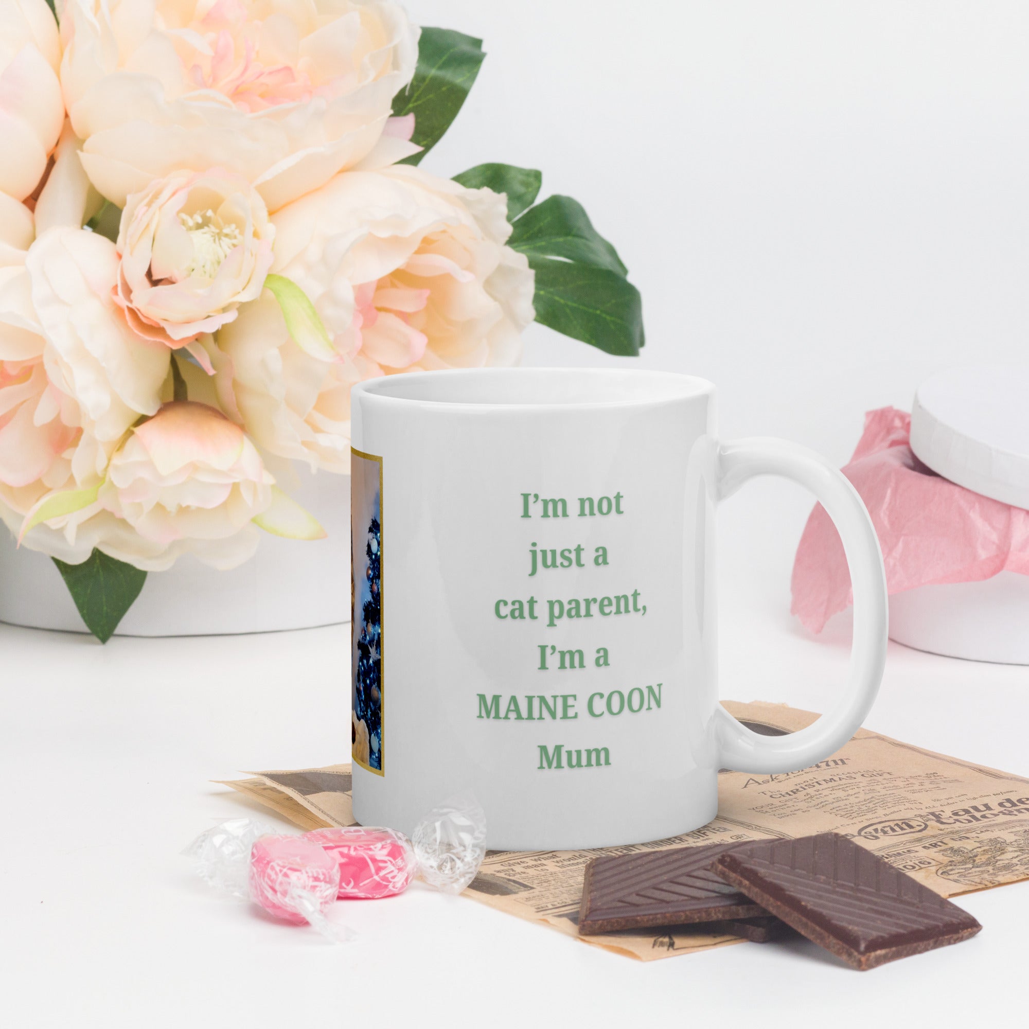 White mug with text 'I'm not just a cat parent, I'm a MAINE COON Mum' next to flowers and chocolate.