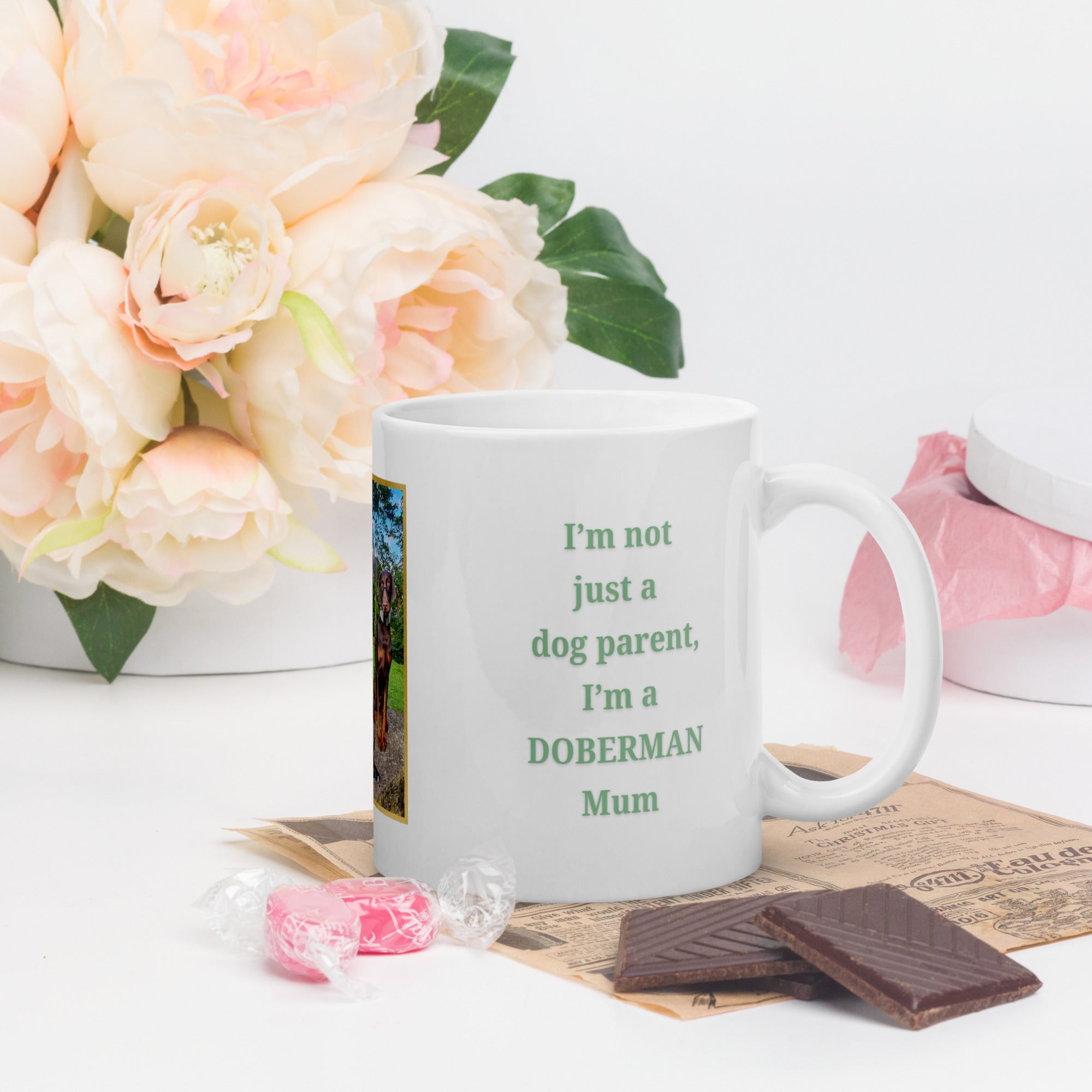 White mug with text 'I'm not just a dog parent, I'm a DOBERMAN Mum' on a white surface with flowers and chocolate.