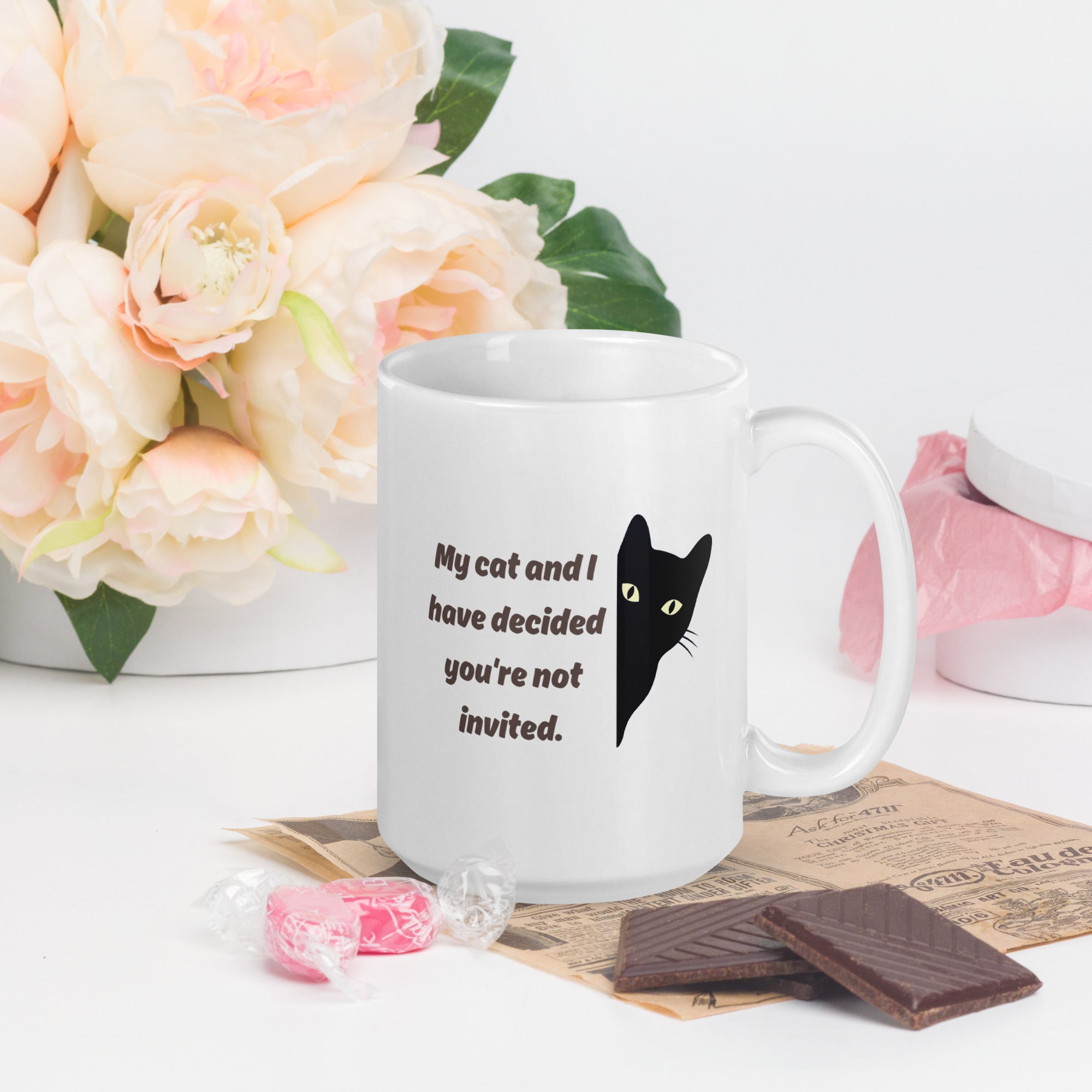 White mug with a black cat illustration and text, placed on a surface with flowers and snacks.